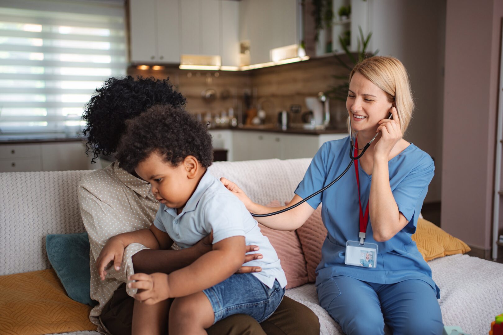 Female pediatrician in blue scrubs using stethoscope to examine young child while comforting and consulting with parents during family home care visit