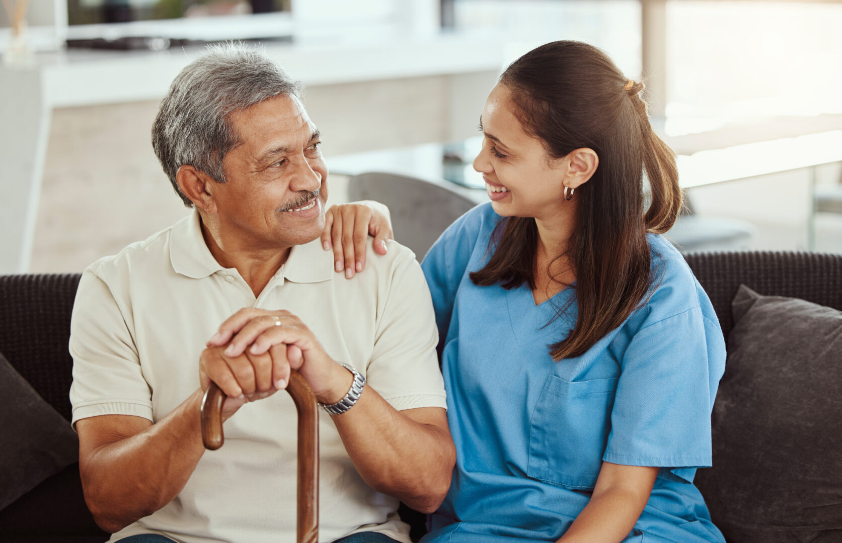 Female nurse in blue scrubs supporting smiling senior man holding cane during medical assistance and rehabilitation therapy session