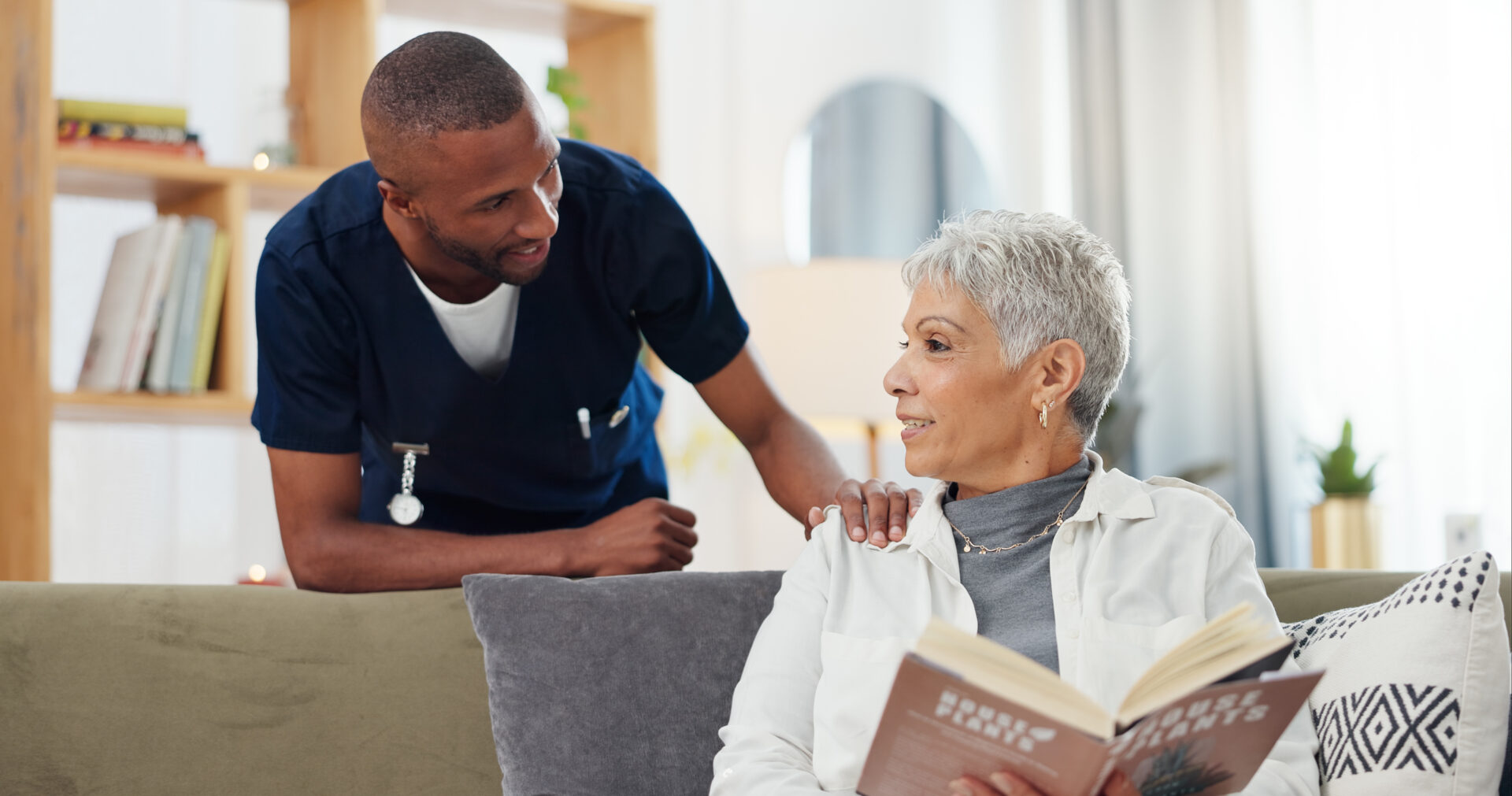 Male nurse in navy scrubs standing beside senior woman reading book on sofa during supportive conversation and literacy engagement at home