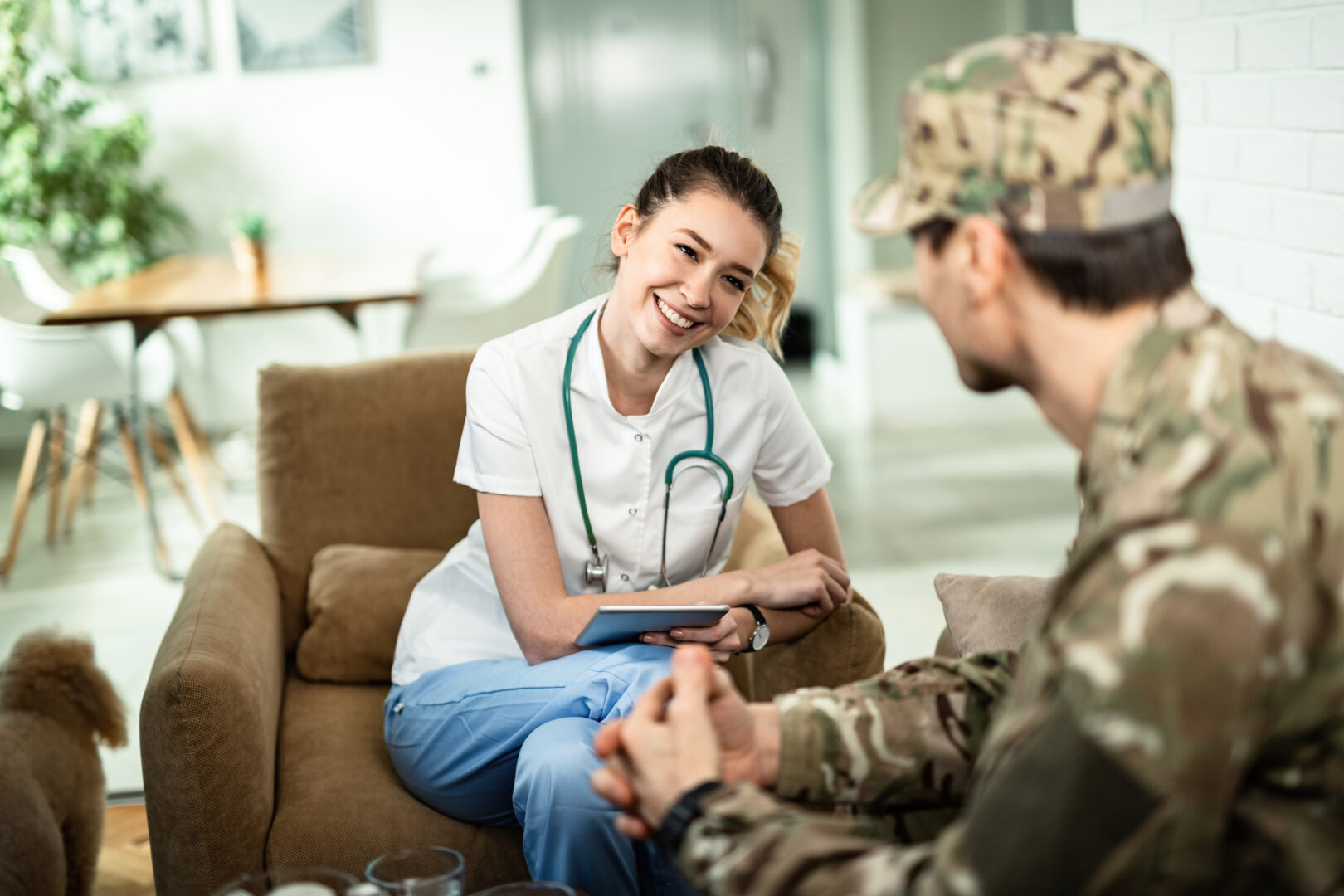 Smiling female healthcare provider with stethoscope consulting with military serviceman holding digital tablet during home care consultation